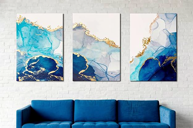 Tres cuadros abstractos azules con detalles dorados colgados sobre un sofá azul en salón moderno